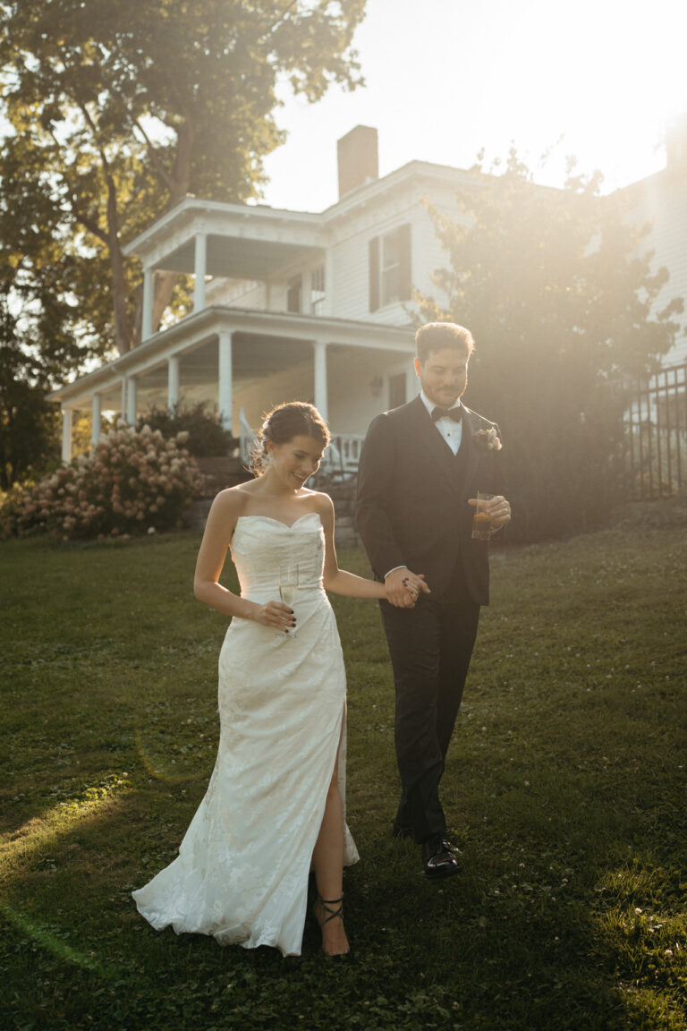 Couple holding hands and walking across a lawn toward a white mansion at sunset