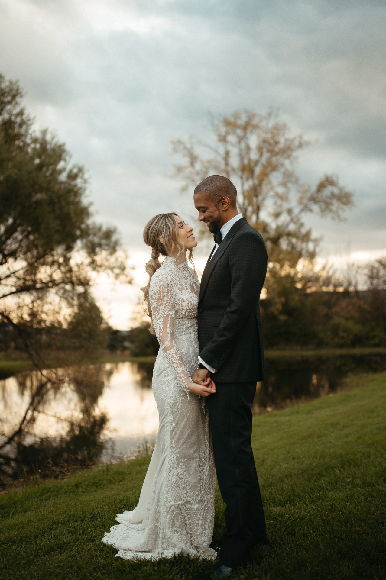 Couple in a lace gown and tuxedo holding hands beside a pond at dusk