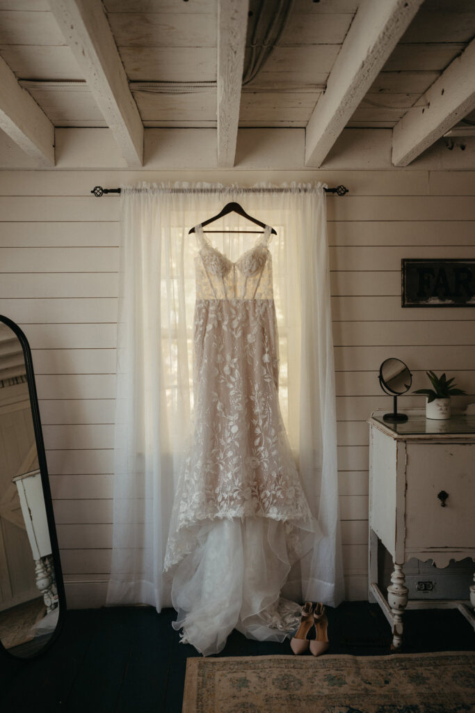Wedding gown hanging in front of sheer curtains in a white-paneled room