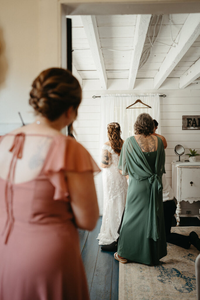 Two attendants in pink and green gowns helping with a wedding gown in a white room
