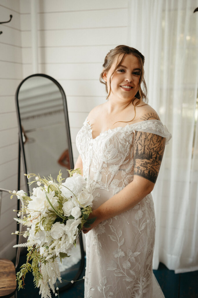 Person in an off-the-shoulder lace gown smiling and holding a white bouquet