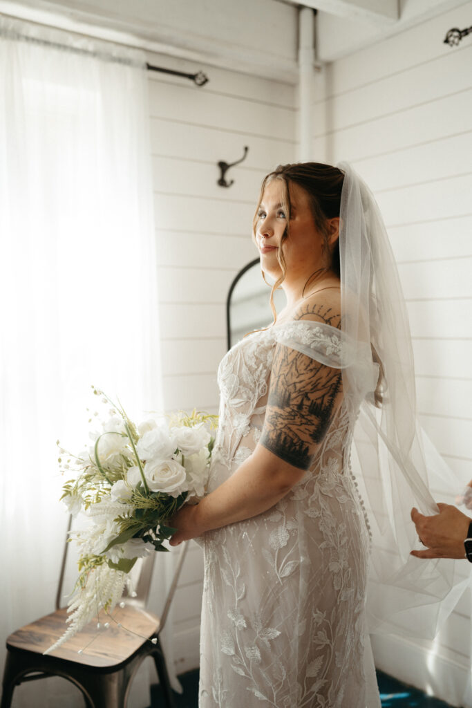 Person in a lace gown and veil holding a bouquet, looking out a window