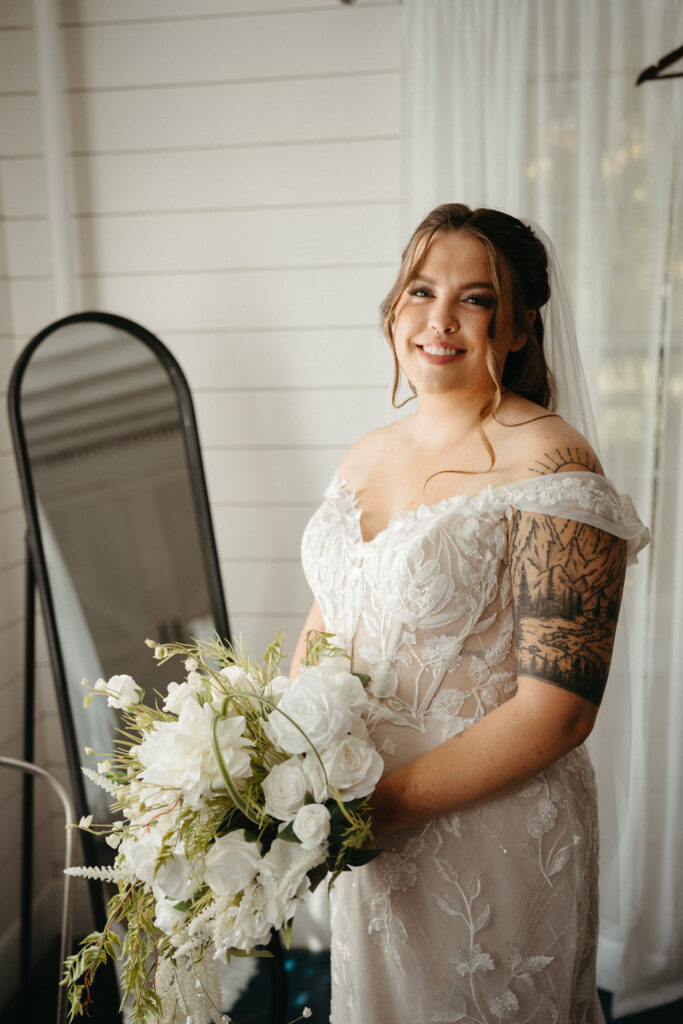 Person in a lace gown and veil smiling, holding a white bouquet beside a mirror