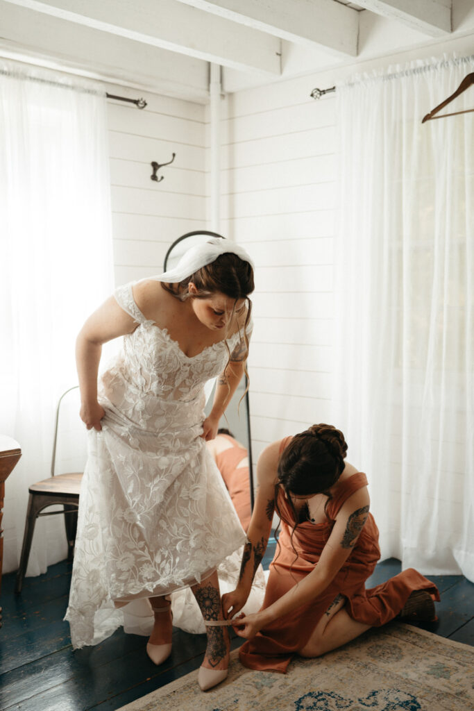 Person adjusting a lace gown while an attendant kneels to help with their shoes