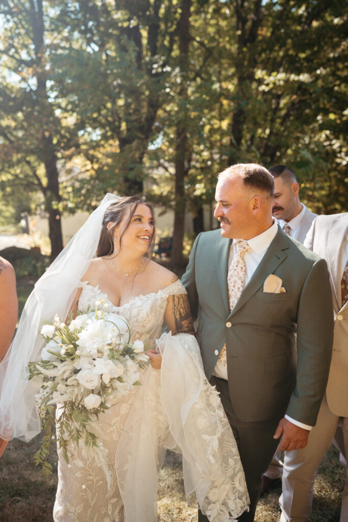 Person in a veil smiling beside a person in a green suit, wedding party behind them