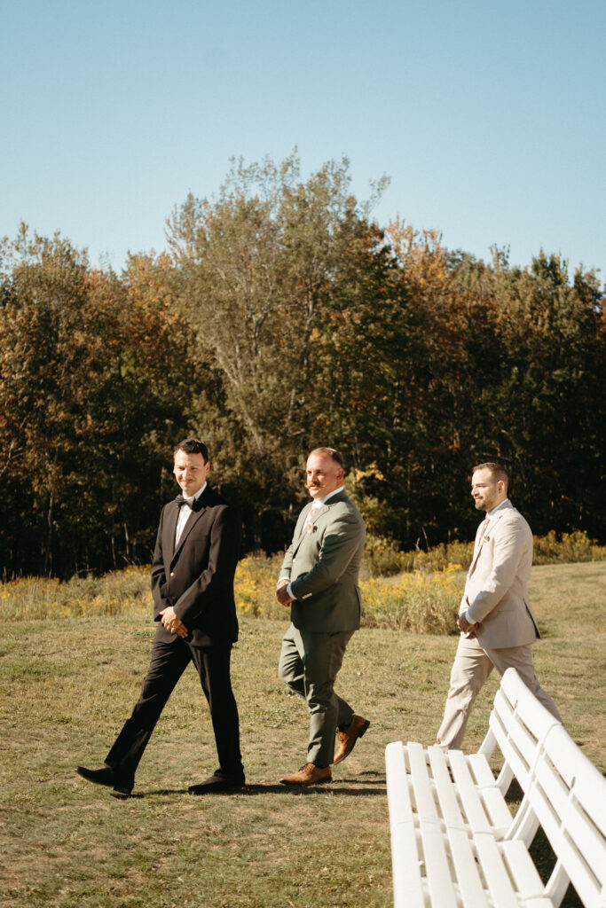 Three people in formal wear walking toward white benches in a meadow