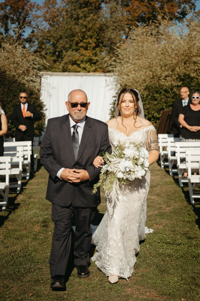 Person in a dark suit escorting a person in a lace gown down a grass aisle