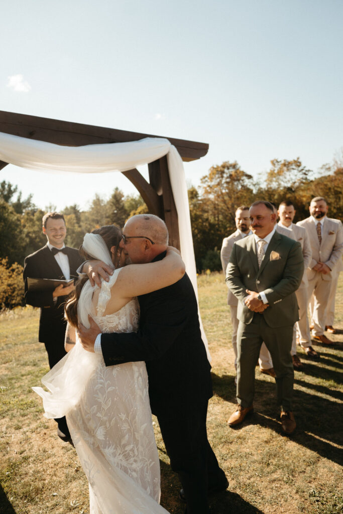 Two people embracing at the ceremony arch while the wedding party looks on