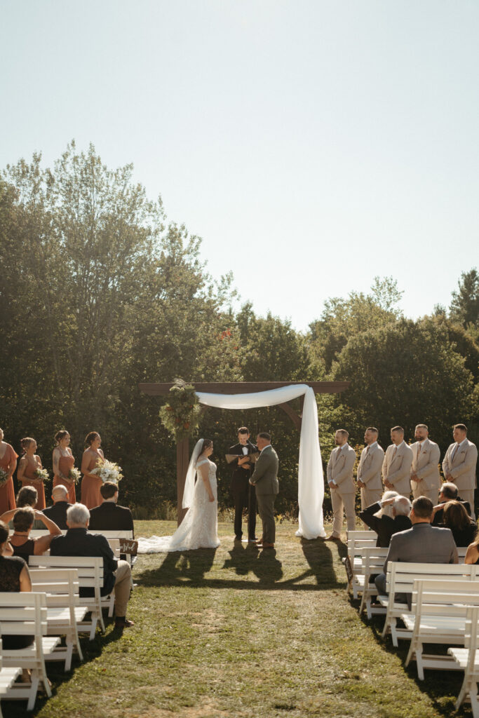 Ceremony under a draped arch with the wedding party and guests on white benches