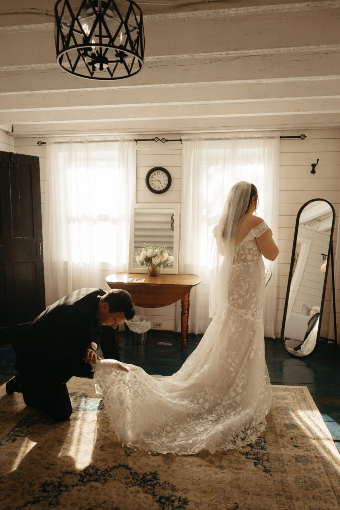 Person in a green suit kneeling to arrange the train of a lace gown in a white room