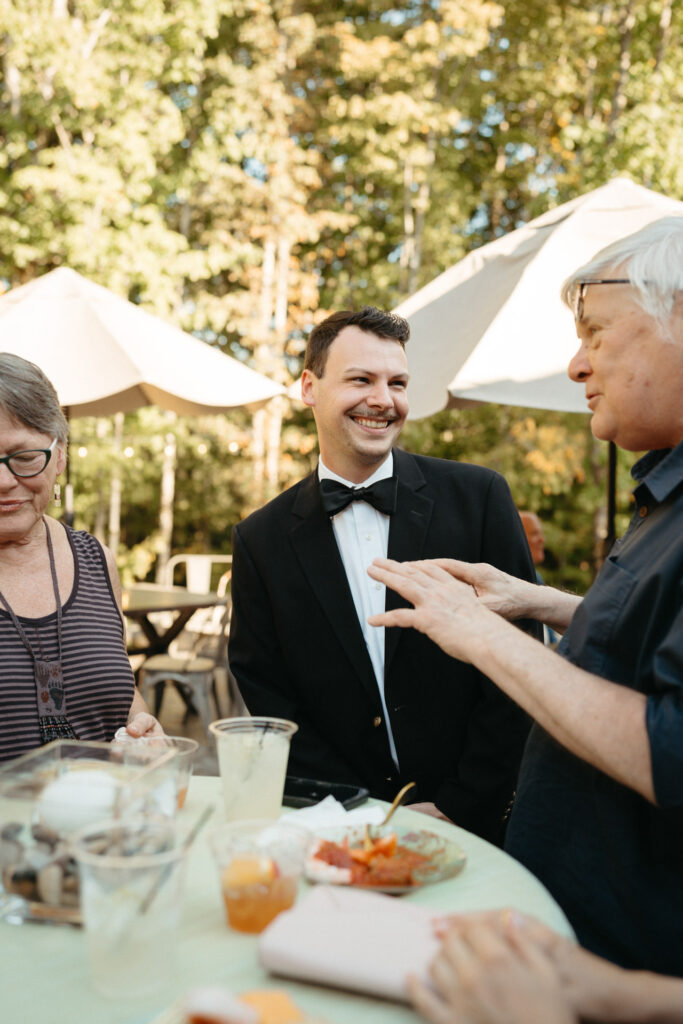 Smiling person in a bow tie chatting with guests at a table under umbrellas