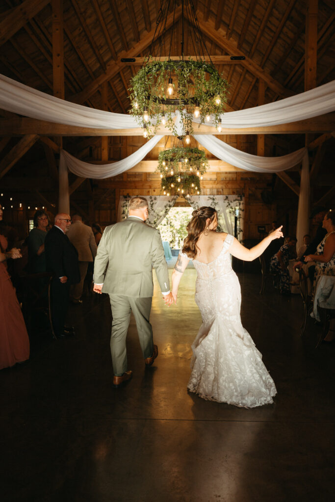 Couple walking onto the dance floor in a barn with draped fabric and chandeliers