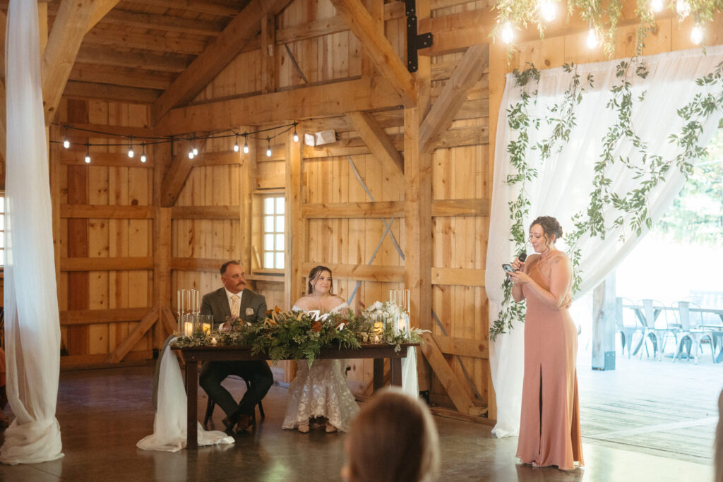 Couple seated at a head table in the barn while a guest gives a toast