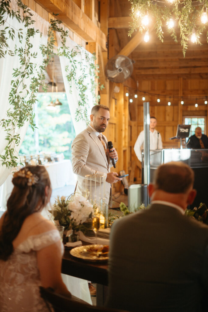 Person in a beige suit giving a toast with the couple seated at the head table