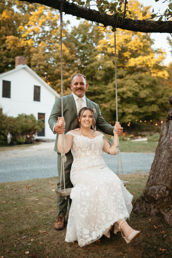 Couple on a tree swing beside a white farmhouse, one seated and one standing
