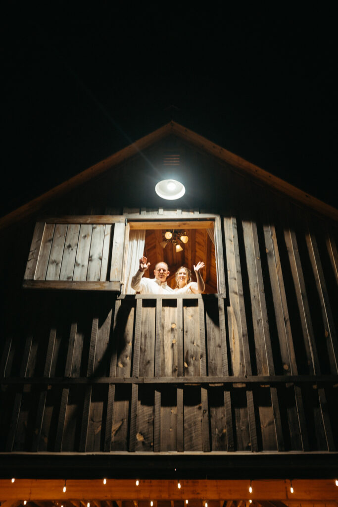 Two guests waving from a hayloft window of the barn at night
