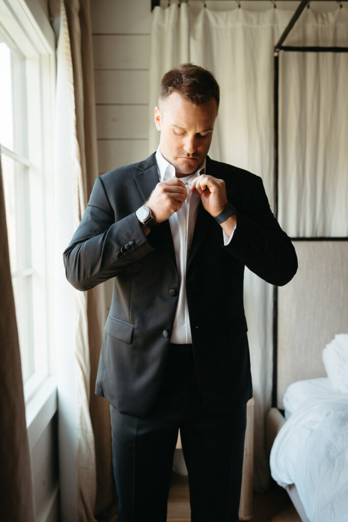 Person in a dark suit buttoning their shirt by a window