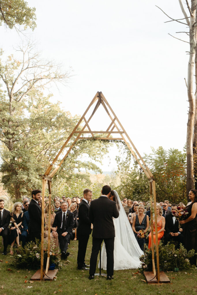 Couple under a wooden A-frame arch with guests seated around them