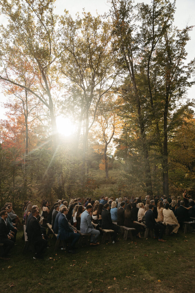 Sun flaring through autumn trees above seated ceremony guests