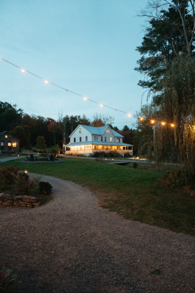 Farmhouse lit up at dusk behind a gravel path and string lights