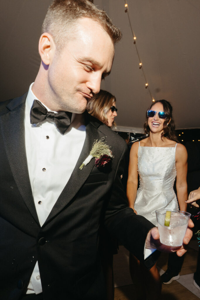 Person in a tuxedo smiling beside a person in a white dress holding a drink