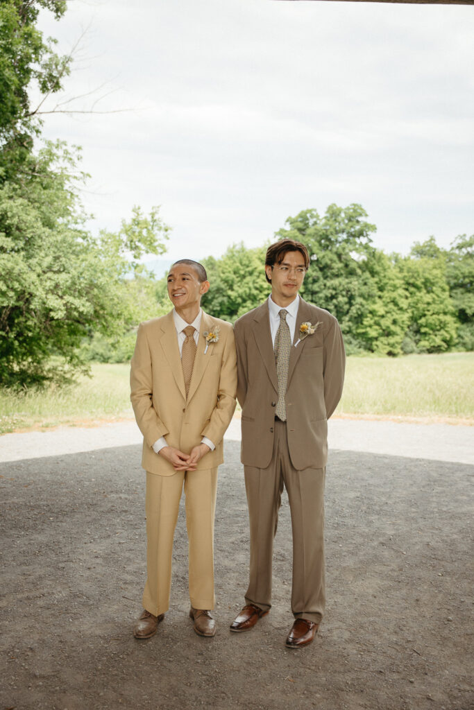 Two people in tan and taupe suits standing on gravel in front of trees