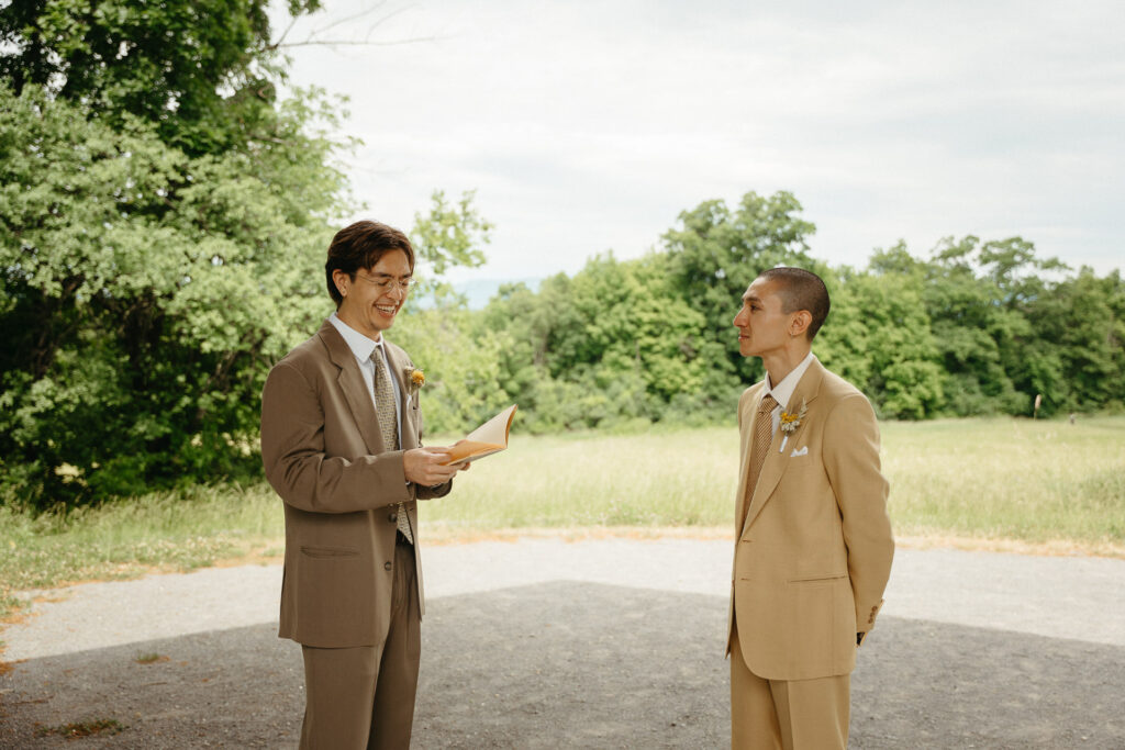 Person in a taupe suit reading vows to the person in a tan suit in front of the field
