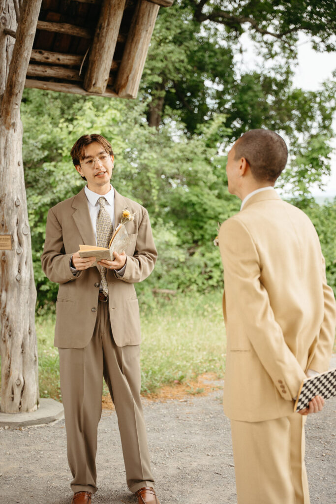 Person in a taupe suit reading vows while the person in a tan suit holds a checkered notebook