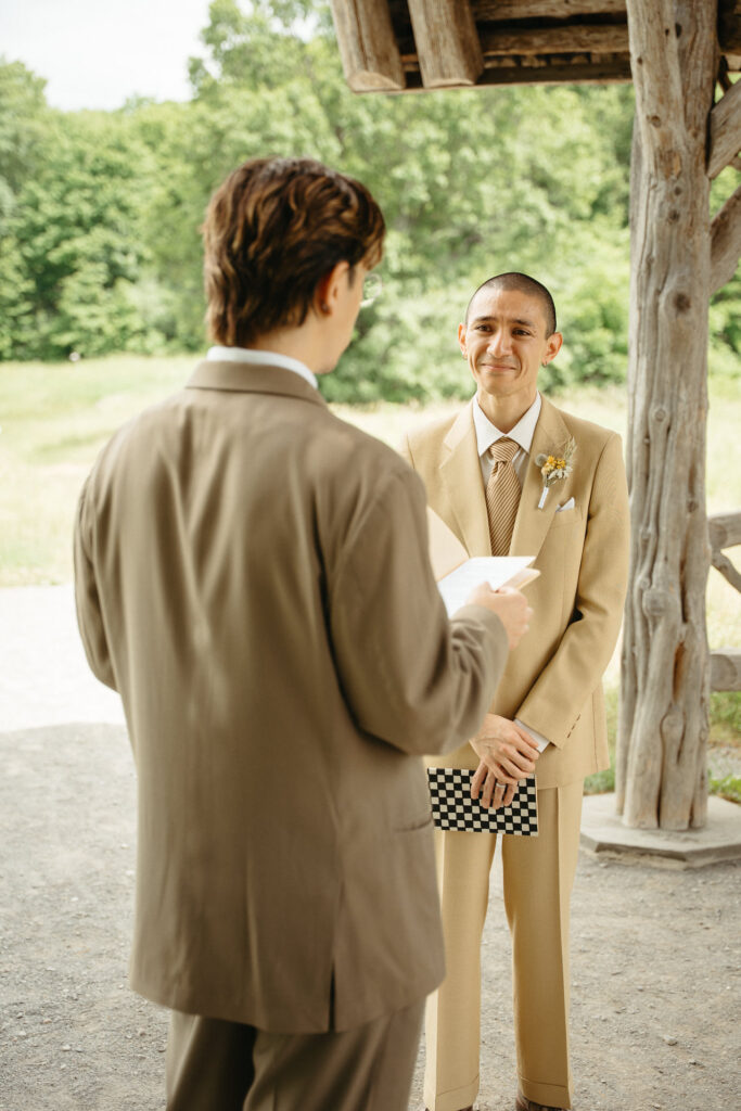 Person in a taupe suit reading as the person in a tan suit smiles, a checkered notebook behind their back