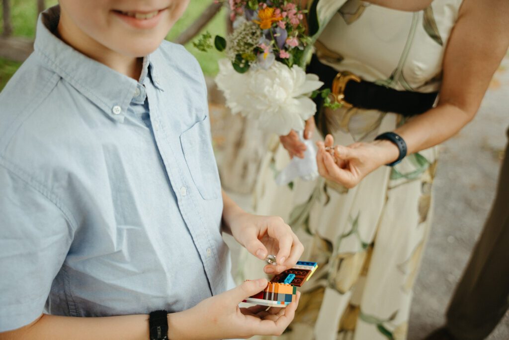 Child in a blue shirt holding a small ring box beside a guest in a floral dress with a bouquet