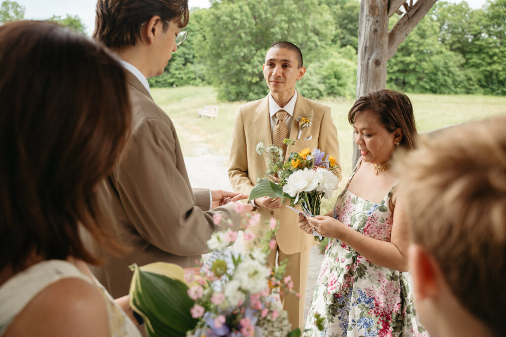 Person in a tan suit smiling as a guest in a floral dress hands over a ring
