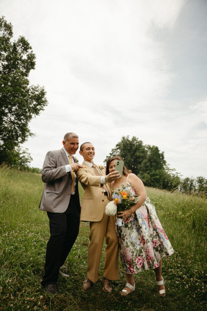 Guest in a gray suit, a person in a tan suit and a guest in a floral dress taking a selfie in the field
