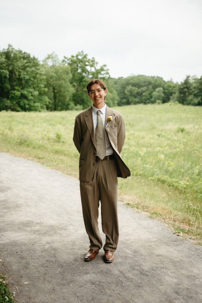 Person in a taupe suit standing on a gravel path with a hand in a pocket